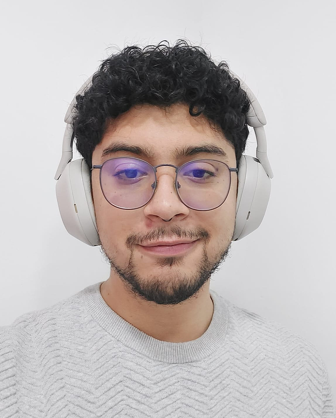 Retrato de Allison, usando audífonos grandes y gafas redondas, sonriendo levemente frente a un fondo blanco.